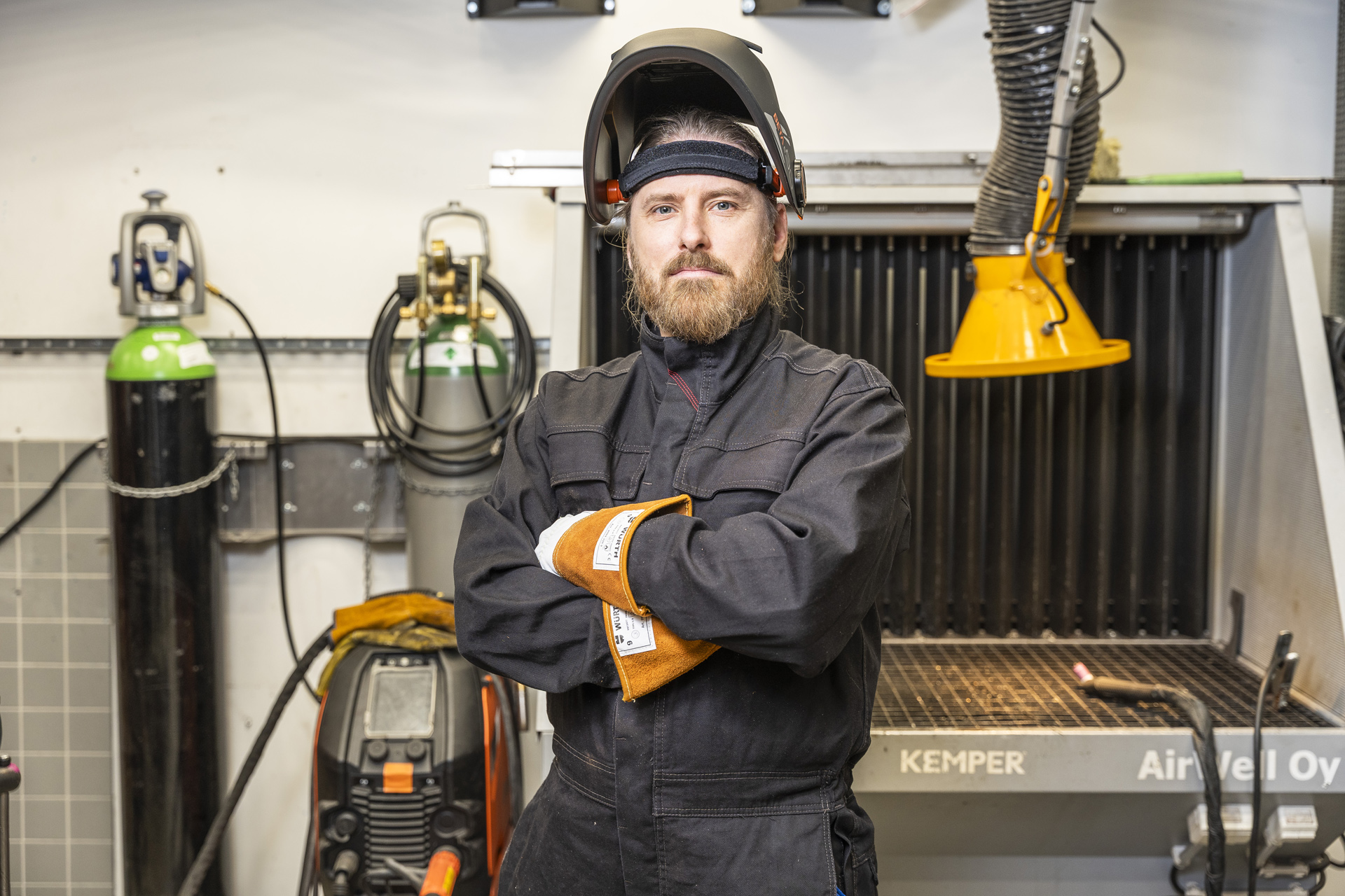 Hitsaussuojavarusteisiin pukeutunut mies seisoo kädet ristissä työpajassa, jonka taustalla on hitsauslaitteita, kaasusäiliöitä ja poistohuppu.