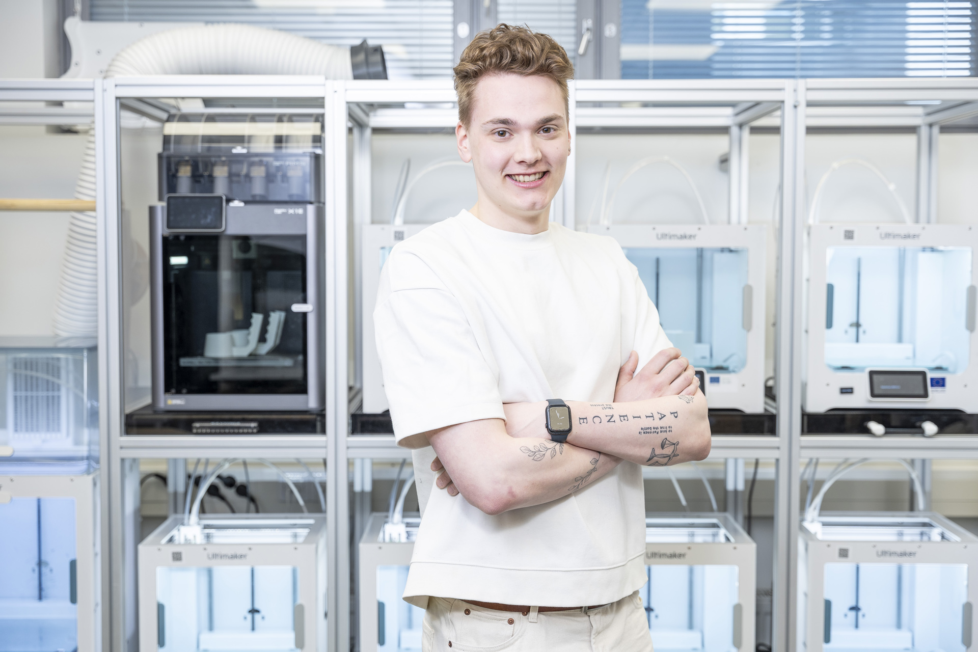 Lyhythiuksinen henkilö, jolla on valkoinen paita ja älykello, seisoo hymyillen ja kädet ristissä useiden 3D-tulostimien edessä valoisassa, modernissa laboratoriossa. Henkilön kyynärvarressa on näkyviä tatuointeja.