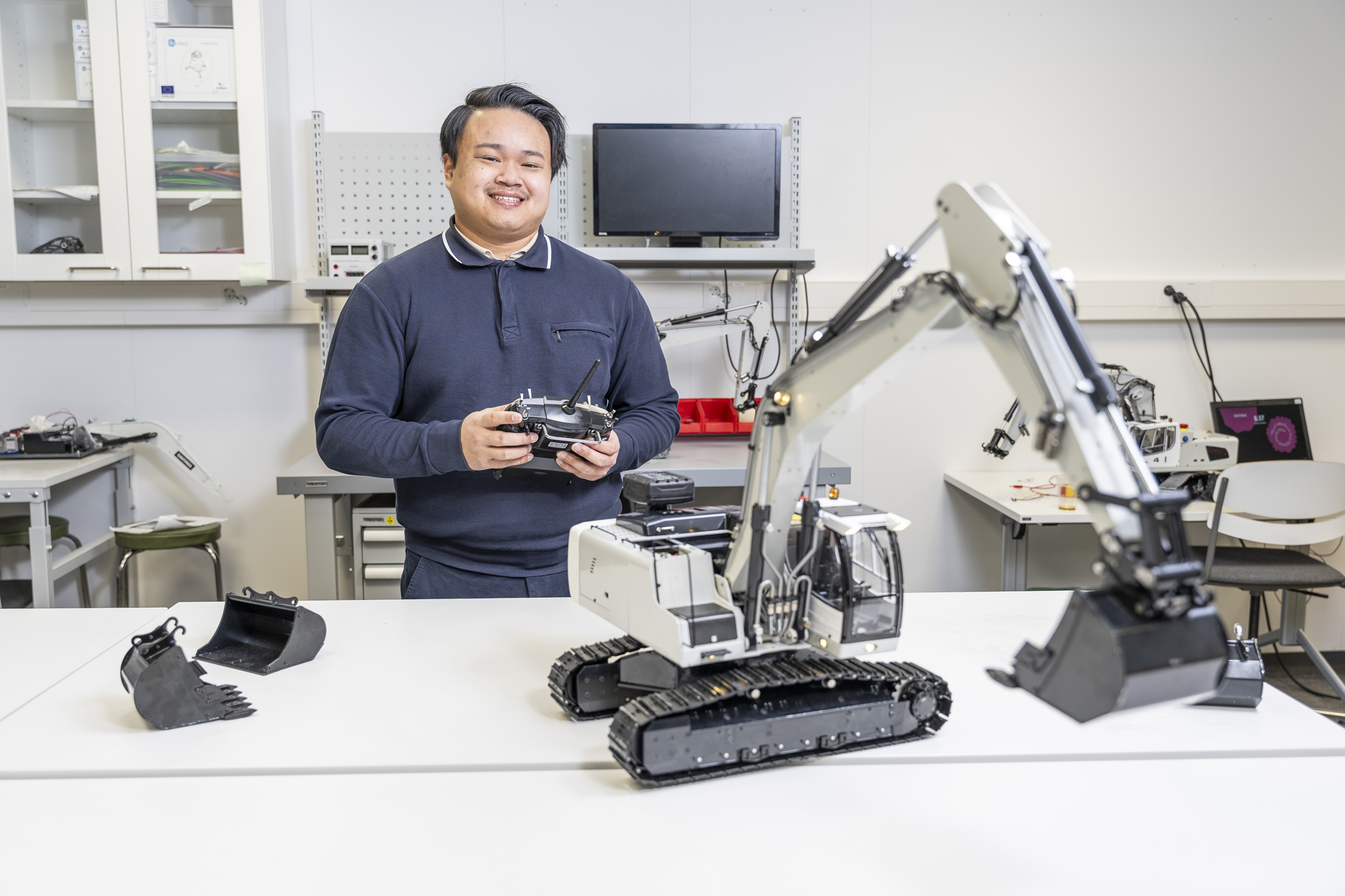 Henkilö seisoo laboratoriossa kaukosäädin kädessään ja hymyilee kameralle. Hänen edessään pöydällä on valkoinen robottikaivinkone ja vaihdettava kaivinkoneen kauha. Taustalla on teknisiä laitteita.