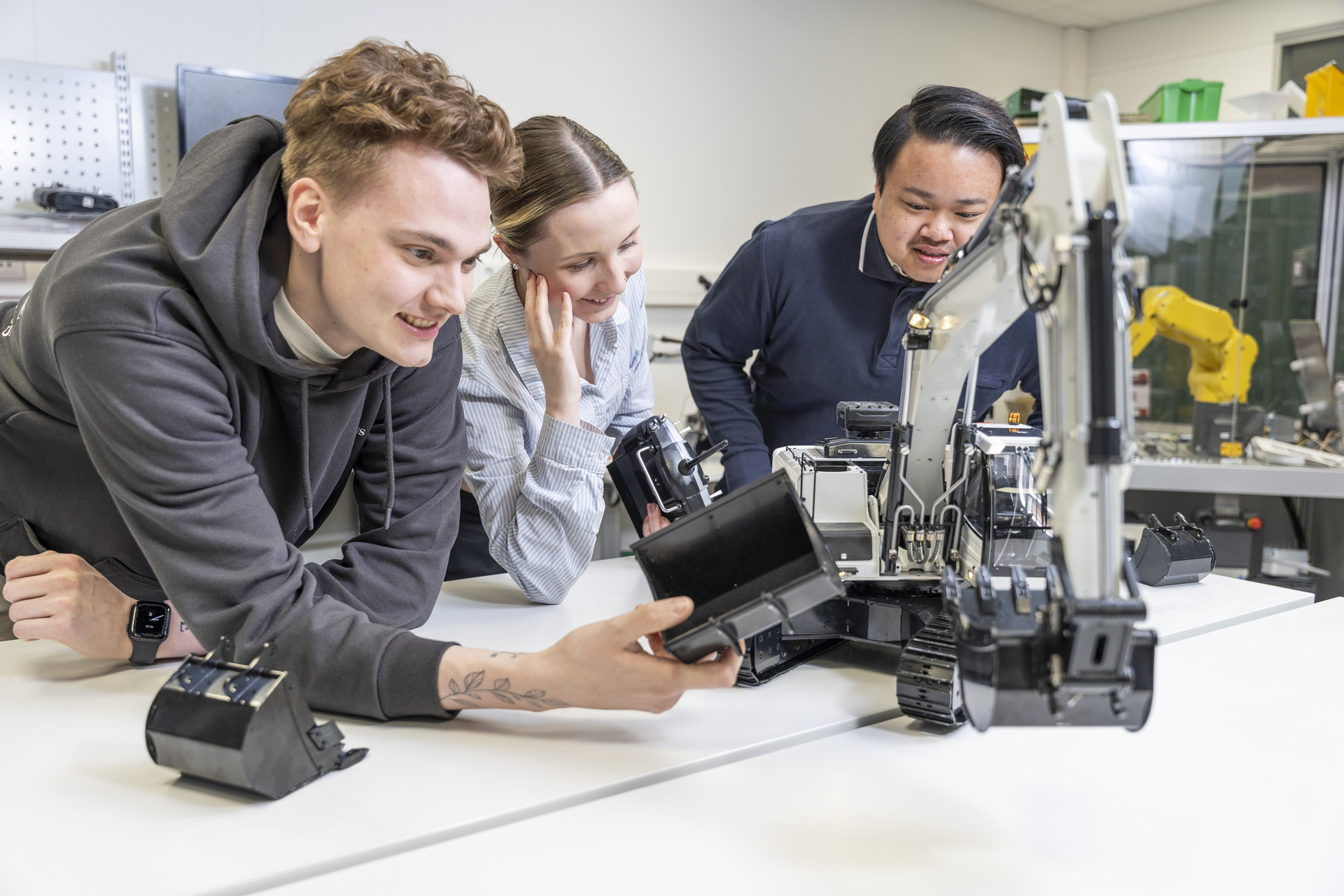 Kolme ihmistä tutkii hymyillen robottikättä valoisassa laboratoriossa pöydällä, vuorovaikutuksessa sen osien kanssa ja keskustelemassa sen ominaisuuksista. Taustalla näkyy erilaisia robotin osia ja laitteita.