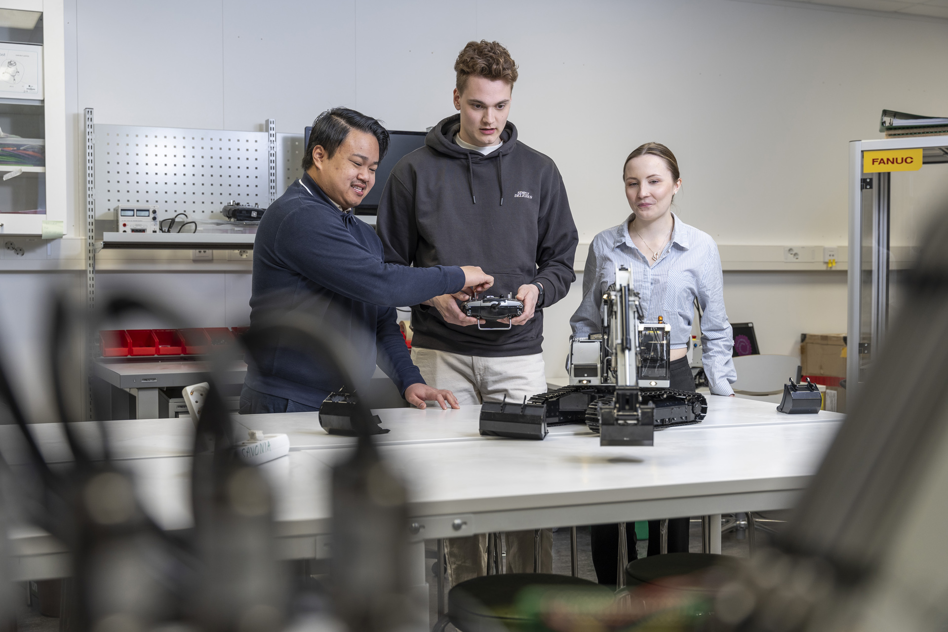 Kolme ihmistä seisoo laboratoriossa pöydän ympärillä ja käyttää ja tarkkailee robottikättä. Yksi henkilö osoittaa robottia, kun muut katsovat tarkkaavaisesti. Taustalla näkyy hyllyjä, työkaluja ja laitteita.