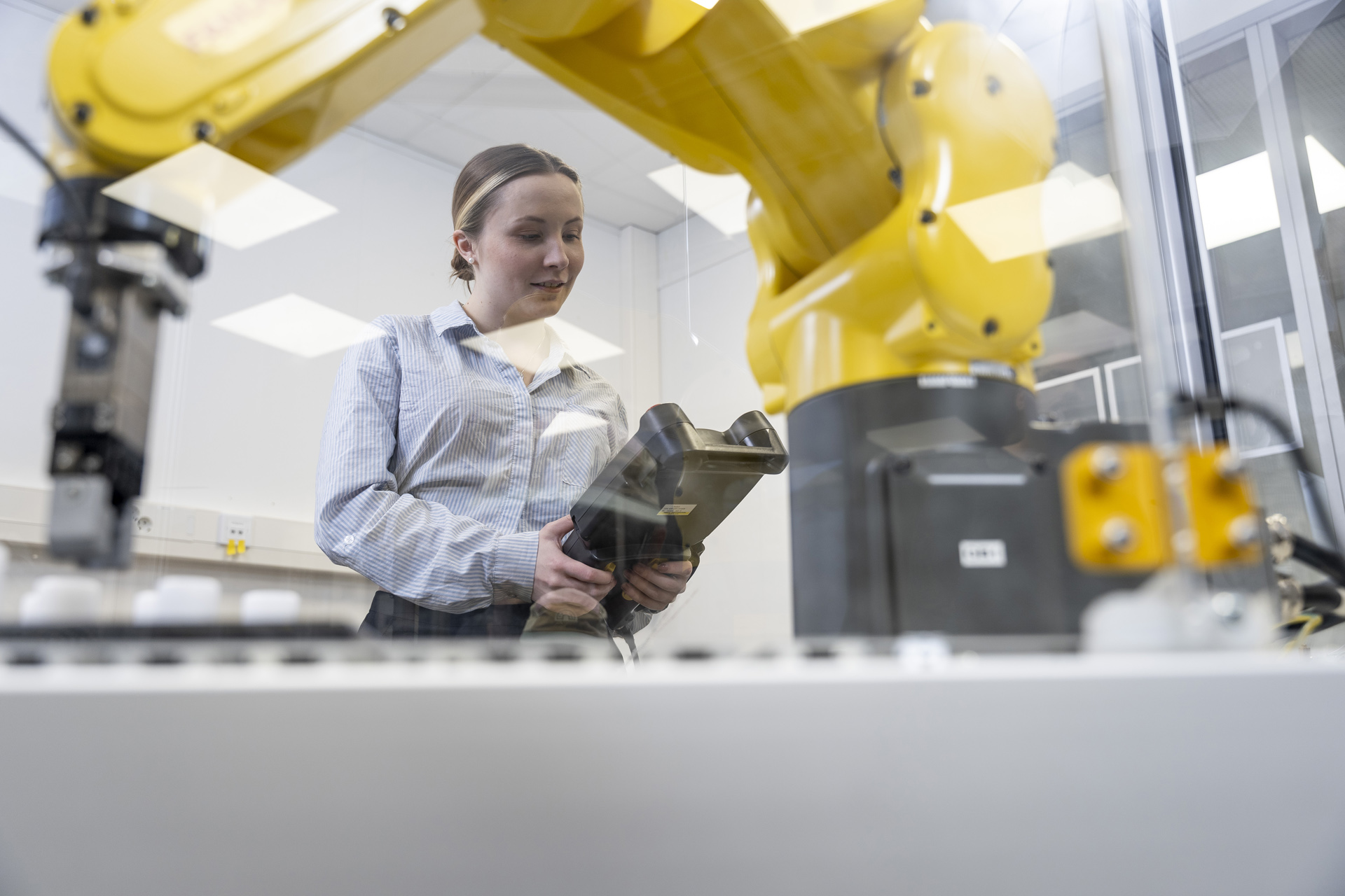 Laboratoriossa työskentelevä nainen käyttää robottikäsivartta käsikäyttöisellä ohjauslaitteella lasipaneelin läpi modernissa laboratoriossa tai teollisuusympäristössä.