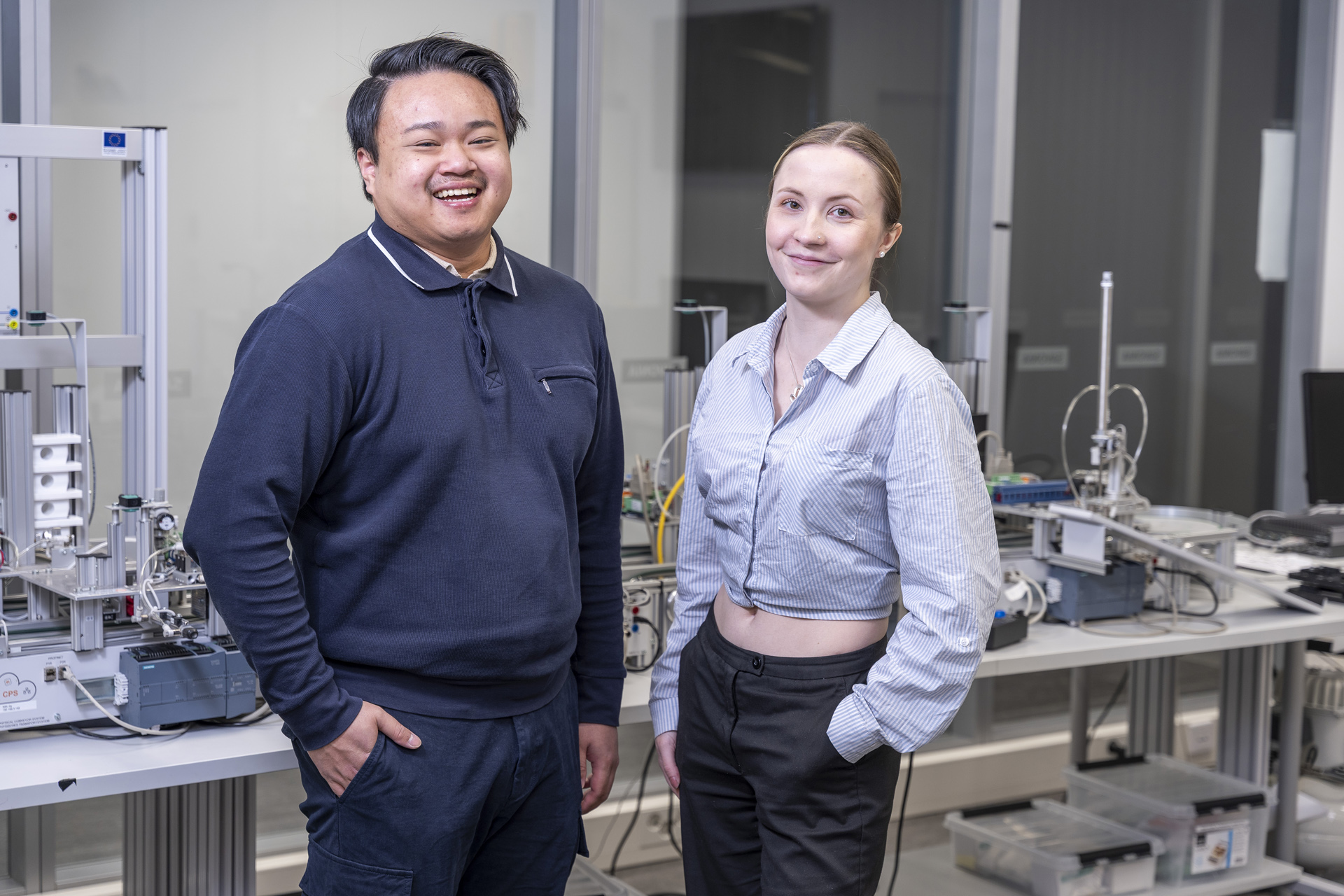 Kaksi hymyilevää ihmistä seisoo modernissa laboratoriossa, jota ympäröivät valkoisilla pöydillä olevat tieteelliset laitteet ja koneet, taustalla lasiseinät ja erilaiset työkalut.