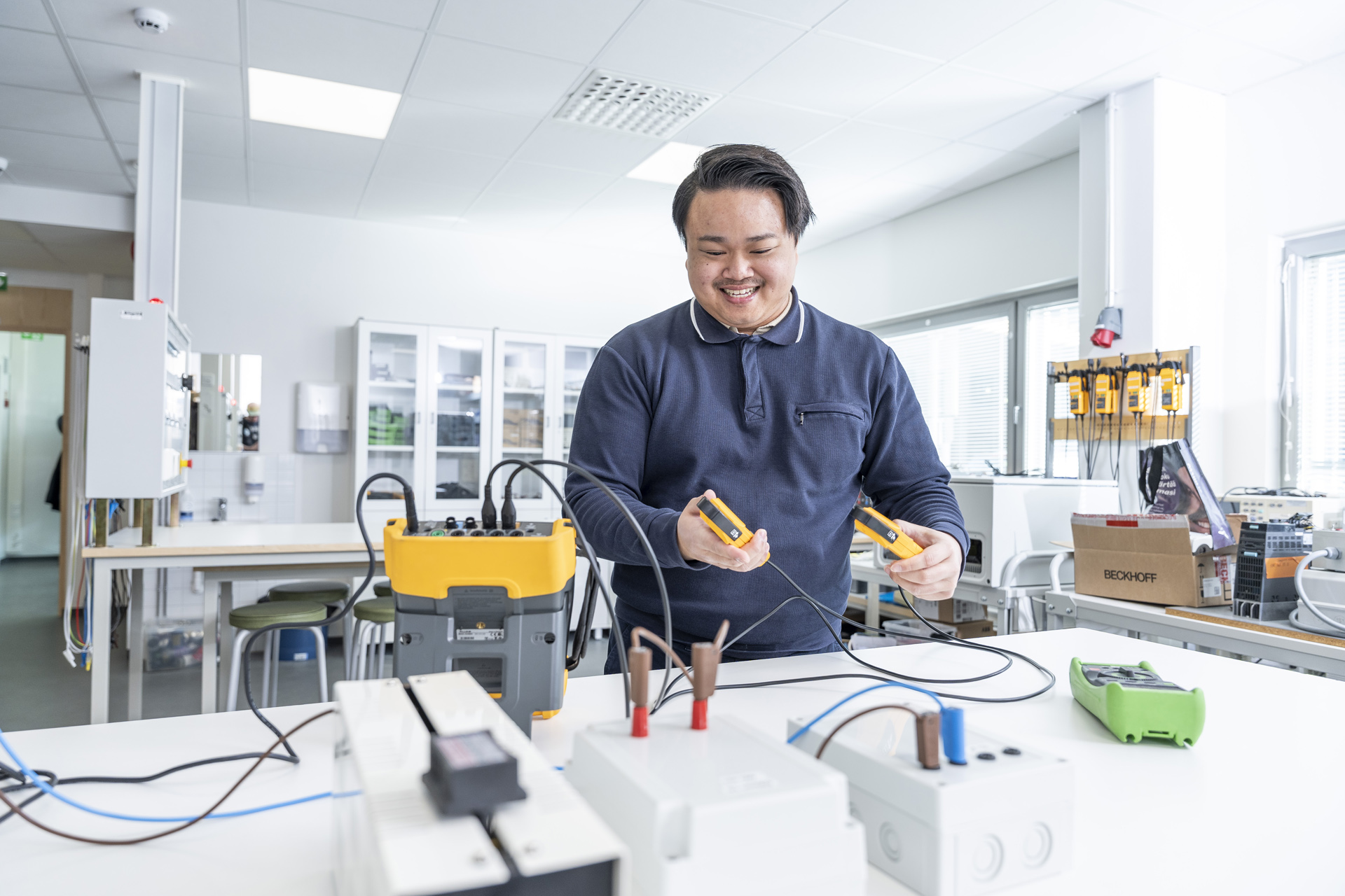 Laboratoriossa työskentelevä henkilö käyttää sähkötestauslaitteita ja hymyilee pitäen kädessään keltaiseen laitteeseen liitettyjä antureita. Työtilassa on erilaisia työkaluja, kaapeleita ja laitteita valkoisilla pöydillä.