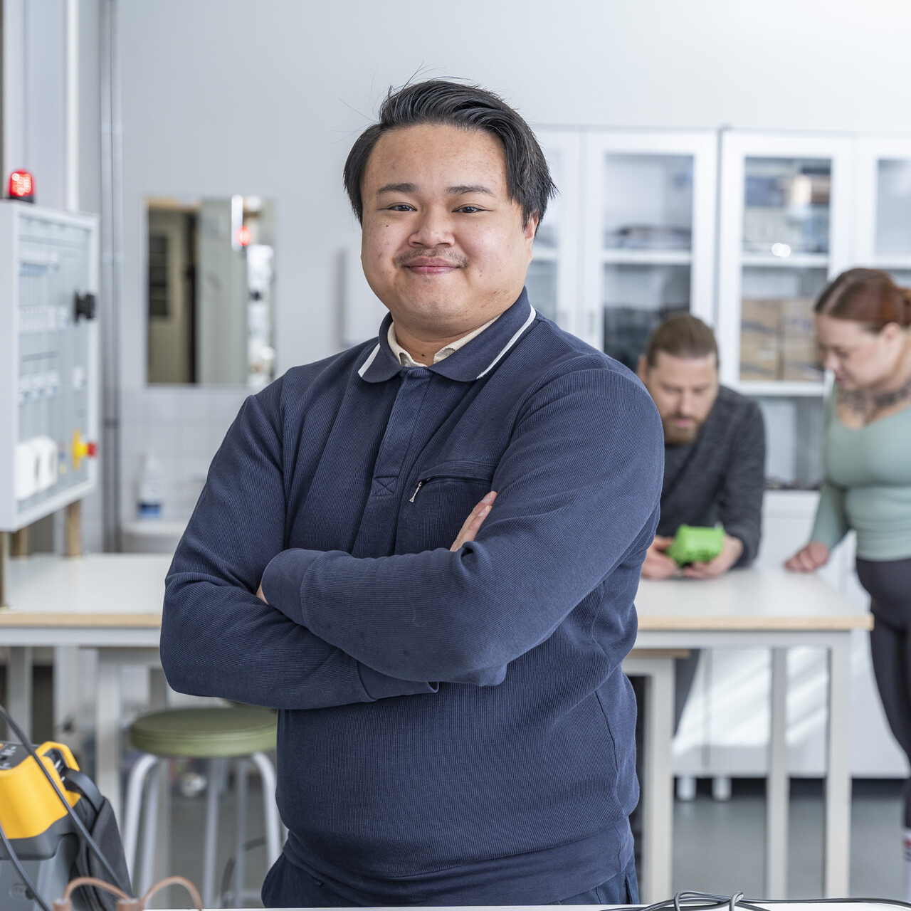 Henkilö seisoo itsevarmasti kädet ristissä valoisassa laboratoriossa, kun taas kaksi muuta työskentelee taustalla pöydän ääressä elektronisten laitteiden kanssa.