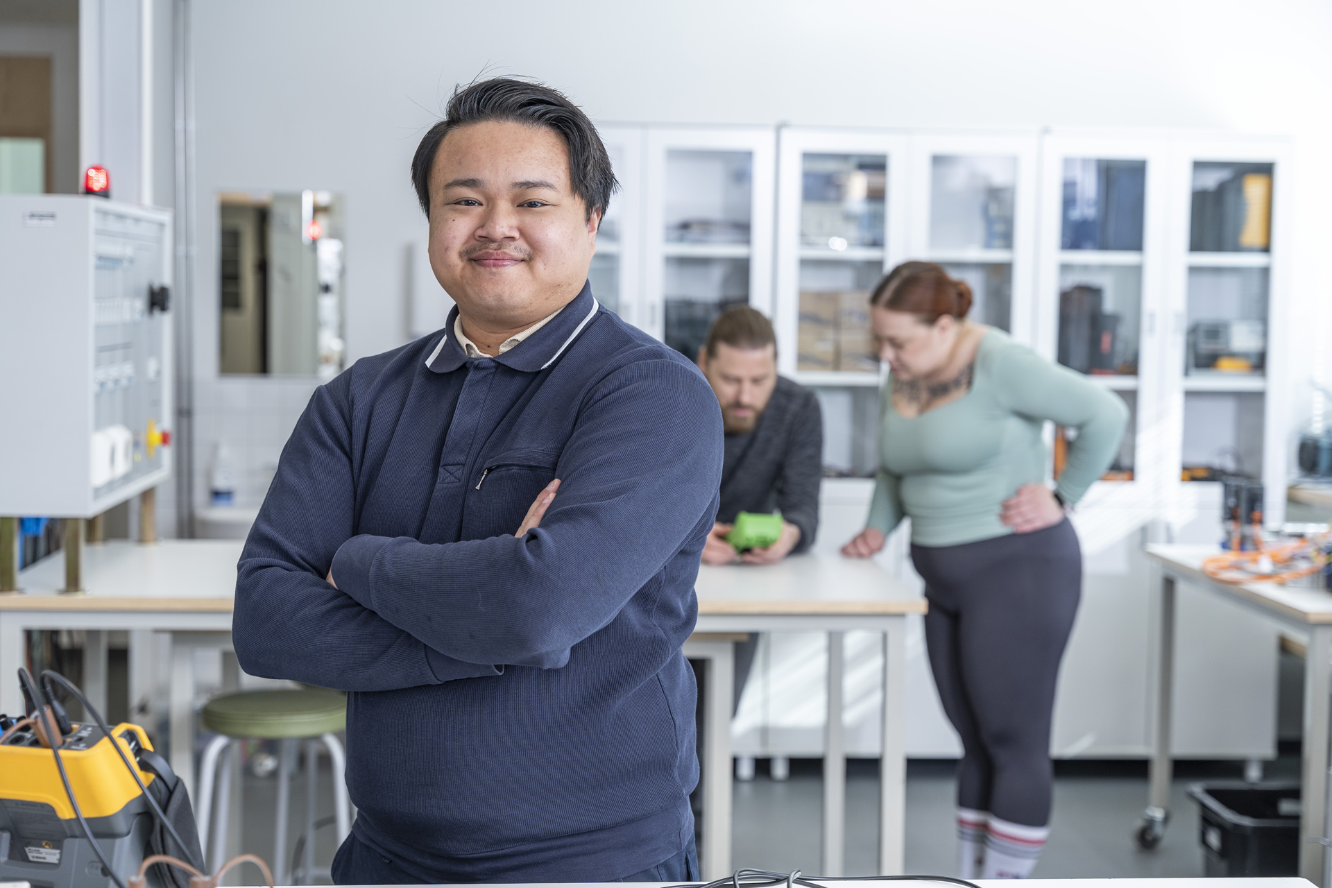 Henkilö seisoo itsevarmasti kädet ristissä valoisassa laboratoriossa, kun taas kaksi muuta työskentelee taustalla pöydän ääressä elektronisten laitteiden kanssa.