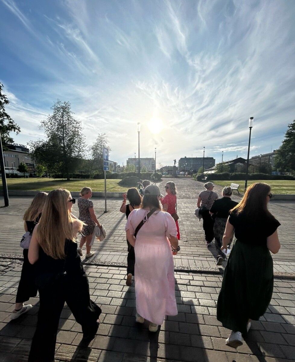 Joukko ihmisiä, enimmäkseen kesämekkoon pukeutuneita naisia, kävelee yhdessä aurinkoisena päivänä pitkin päällystettyä polkua, jonka molemmin puolin on puita ja vihreää ruohoa. Aurinko paistaa kirkkaasti siniseltä taivaalta, jonka pilvet peittävät osittain.