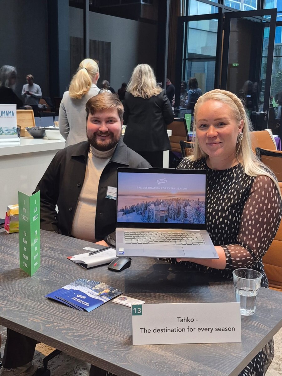Kaksi ihmistä istuu pöydän ääressä sisätiloissa. Oikealla istuva nainen hymyilee ja pitää kädessään kannettavaa tietokonetta, jossa näkyy kuva lumisesta maisemasta. Vasemmalla istuva mies hymyilee myös. Pöydällä olevassa kyltissä lukee: "Tahko - Kohde joka vuodenaikaan.".