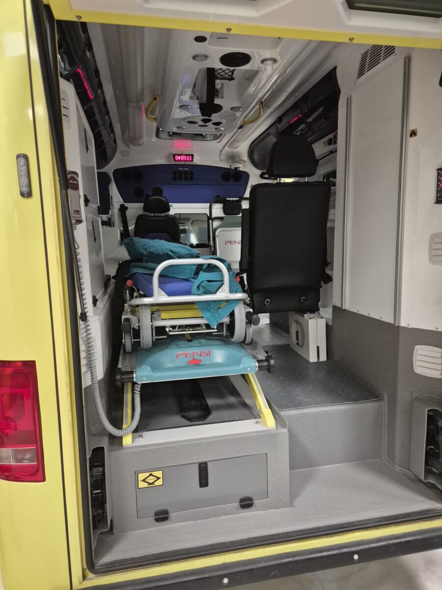 Ambulanssin sisätila, jossa näkyy siniset paarit, lääkinnälliset laitteet ja istuimet. Paarit ovat keskellä, valmiina potilaan kuljetukseen. Tila vaikuttaa siistiltä ja hyvin organisoidulta.