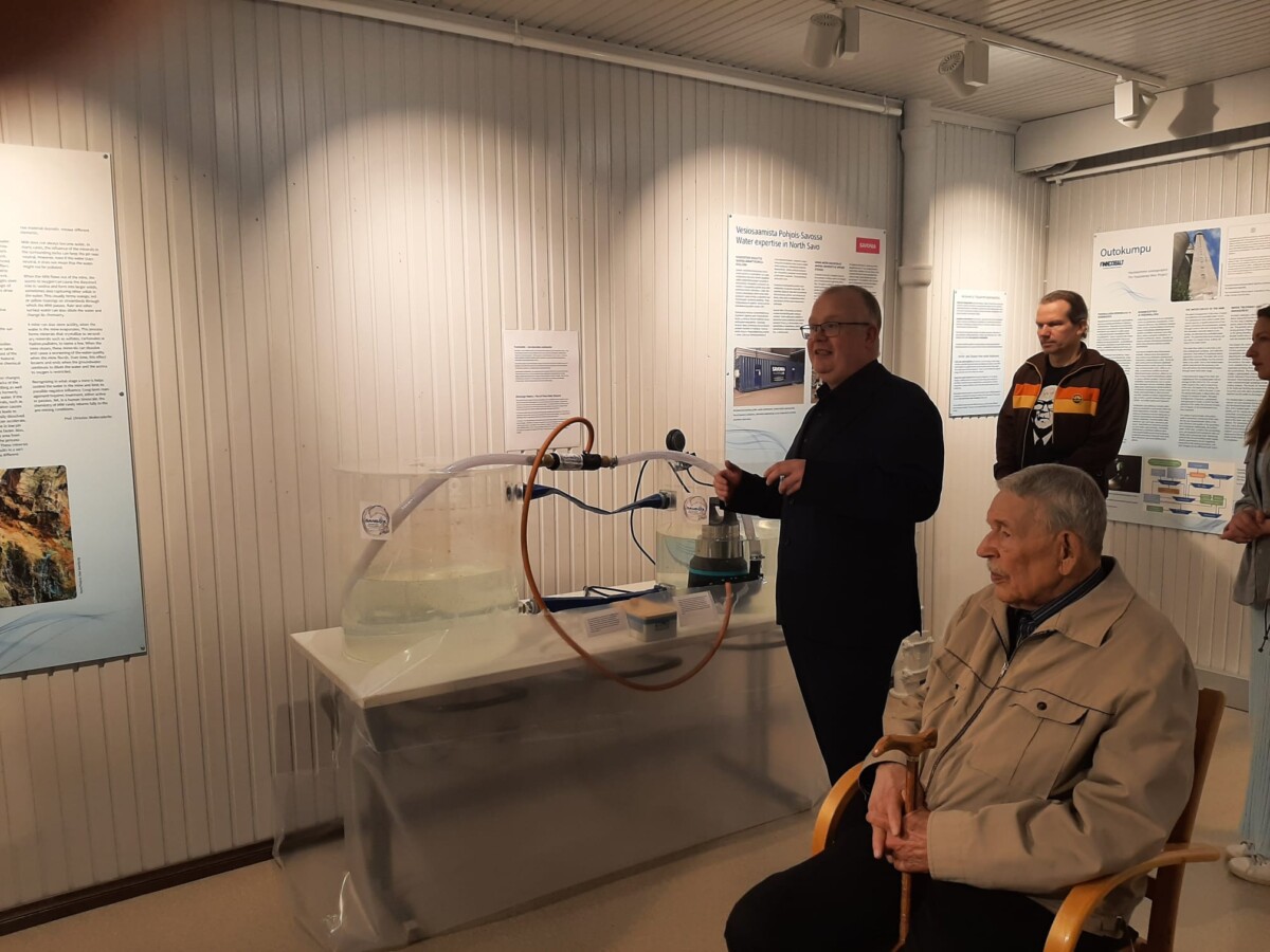 Mies seisoo veden virtausta esittelevän laitteiston vieressä ja puhuu yleisölle museomaisessa näyttelytilassa. Etualalla istuu vanhempi mies, ja taustalla kaksi muuta henkilöä seuraa tilannetta.