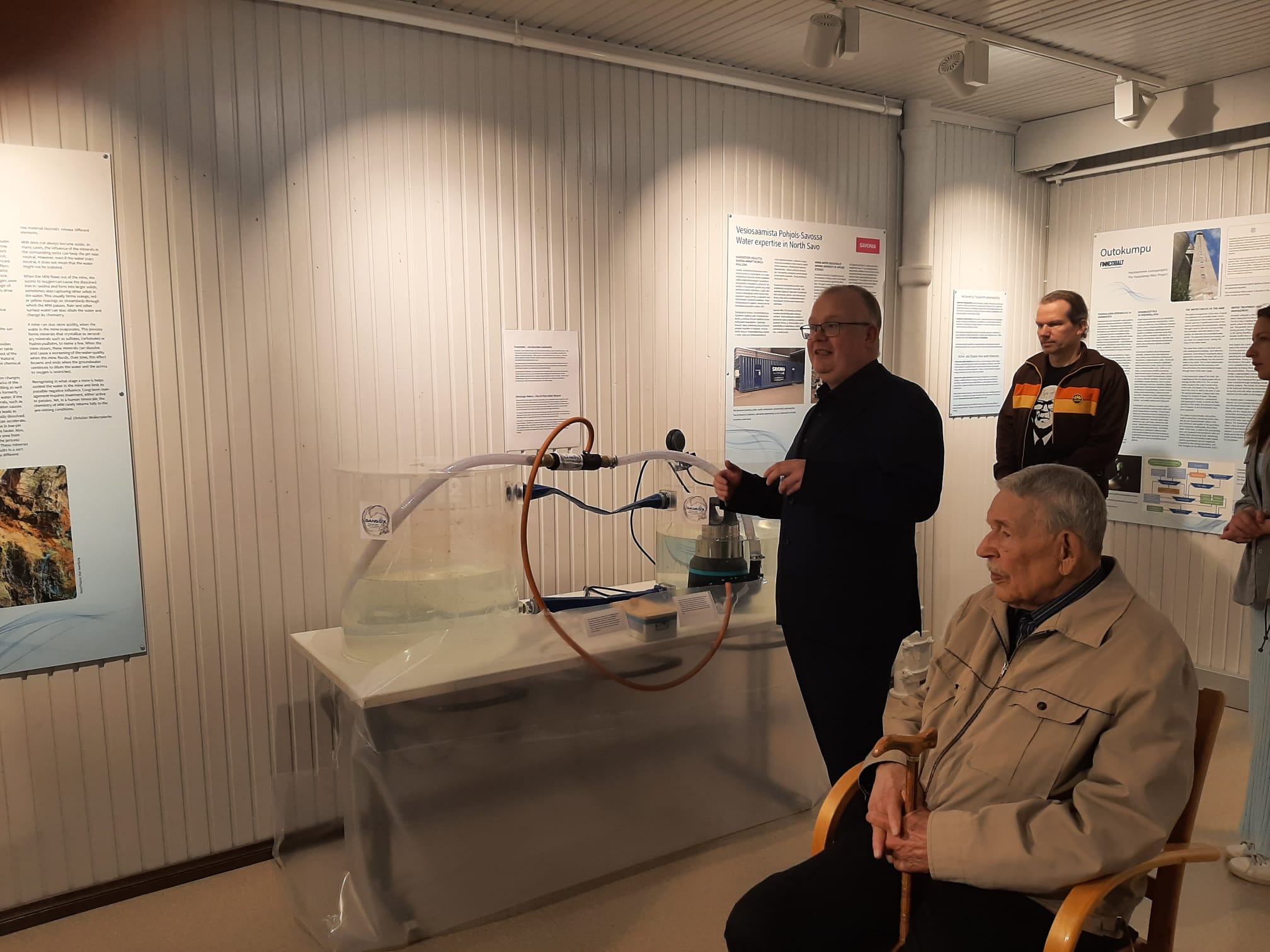 Mies seisoo veden virtausta esittelevän laitteiston vieressä ja puhuu yleisölle museomaisessa näyttelytilassa. Etualalla istuu vanhempi mies, ja taustalla kaksi muuta henkilöä seuraa tilannetta.