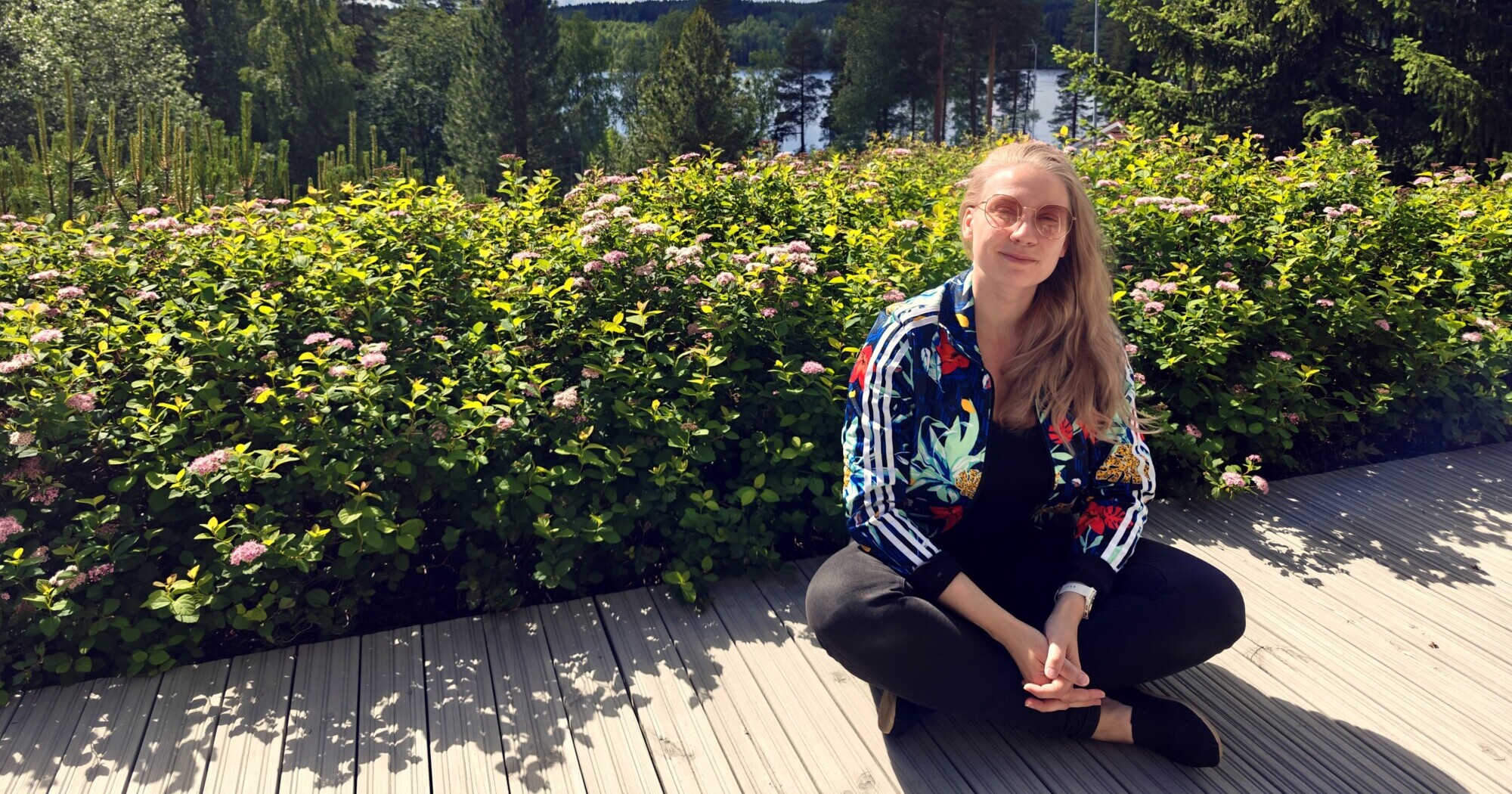 Nainen, jolla on pitkät vaaleat hiukset, silmälasit ja värikäs takki, istuu ristikkäin puuterassilla, ympärillään vihreitä, vaaleanpunaisilla kukilla koristeltuja pensaita, ja taustalla näkyy puita ja järvi.
