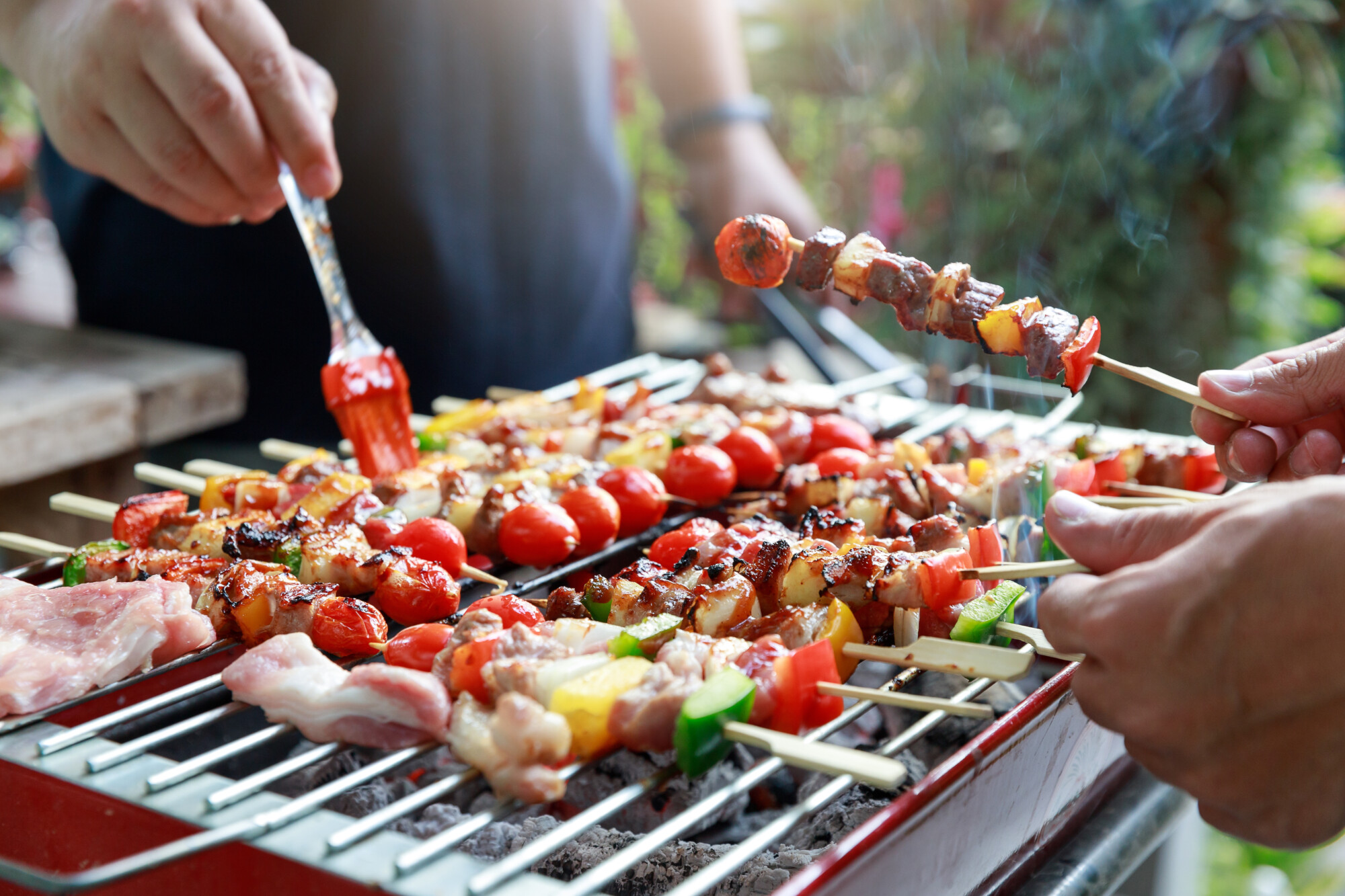 Ihmiset grillata lihavartaita, kirsikkatomaatteja ja vihanneksia grillillä. Yksi henkilö sivelee kastiketta ruoan päälle, kun taas toinen pitää vartta kädessään. Kohtaus viittaa ulkona järjestettävään grillijuhlaan tai grillitapaamiseen.