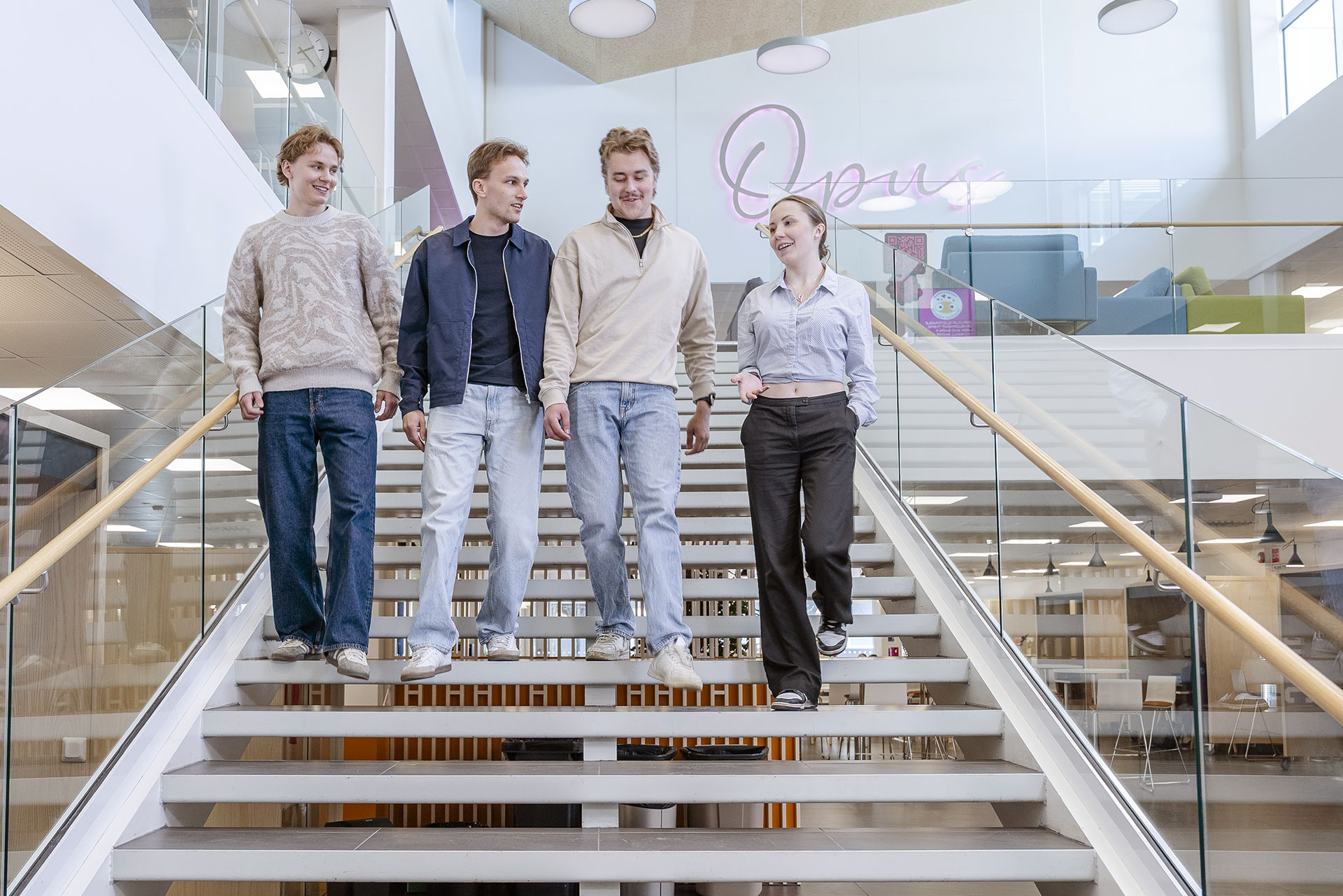 Neljä nuorta aikuista kävelee modernia portaikkoa pitkin sisätiloissa hymyillen ja keskustellen. Taustalla on lasiseinät, neonkyltti, jossa lukee Open, ja valoisa, avoin toimisto- tai opiskeluympäristö.