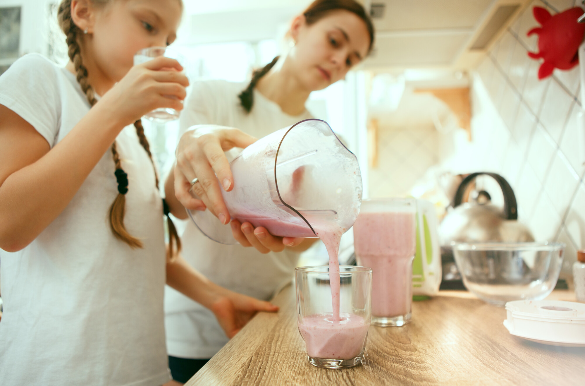 Nainen kaataa vaaleanpunaista smoothieta lasiin, kun nuori tyttö, jolla on punotut hiukset, juo toisesta lasista valoisassa keittiössä.