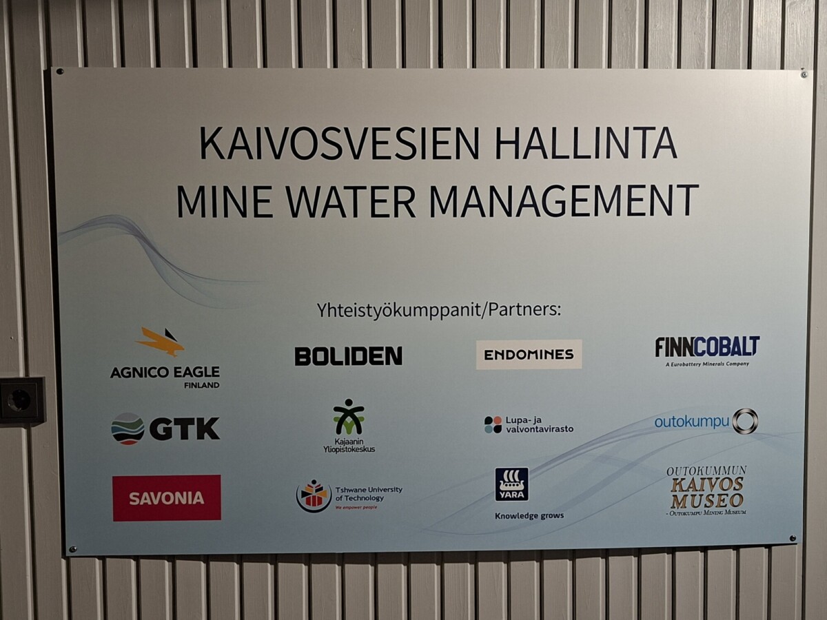 Harmaalle paneeliseinälle on kiinnitetty kyltti, jossa lukee ”KAIVOSVESIEN HALLINTA / MINE WATER MANAGEMENT” ja jossa on luettelo yhteistyöorganisaatioista sekä niiden logot.