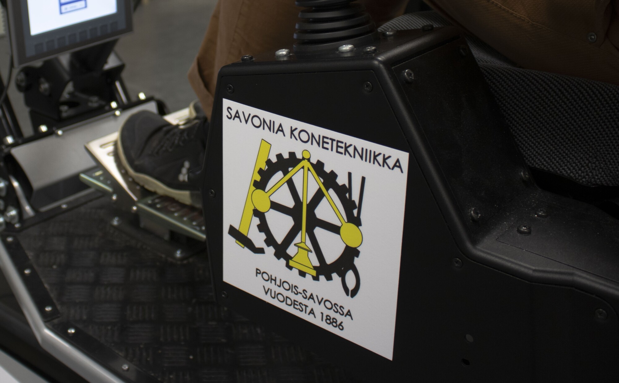 Lähikuva koneesta, jonka etiketissä on kuvattu hammaspyöriä ja työkaluja sekä teksti ”Savonia Konetekniikka, Pohjois-Savossa vuodesta 1886”. Koneen vieressä näkyy osittain henkilön jalka ja kenkä.