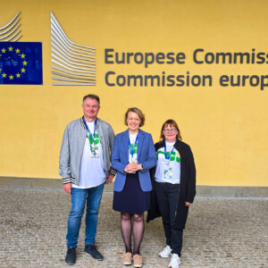 Kolme ihmistä seisoo hymyillen keltaisen seinän edessä, jossa on Euroopan komission logo ja teksti hollanniksi ja ranskaksi. Heillä on yllään tapahtumamerkit ja kaulanauhat.