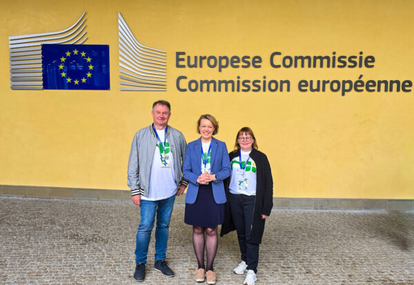 Kolme ihmistä seisoo hymyillen keltaisen seinän edessä, jossa on Euroopan komission logo ja teksti hollanniksi ja ranskaksi. Heillä on yllään tapahtumamerkit ja kaulanauhat.