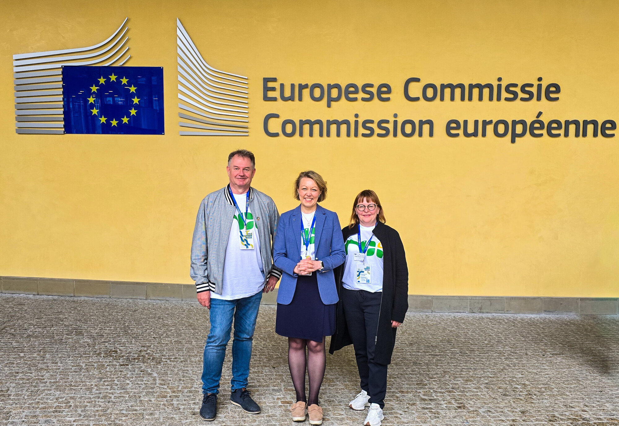 Kolme ihmistä seisoo hymyillen keltaisen seinän edessä, jossa on Euroopan komission logo ja teksti hollanniksi ja ranskaksi. Heillä on yllään tapahtumamerkit ja kaulanauhat.