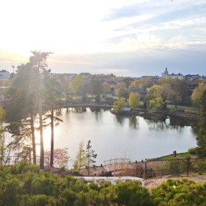Seesteinen lampi puiden ja vehreyden ympäröimänä, auringonvalo paistaa korkeiden puiden läpi. Taustalla näkyy kaupungin rakennuksia ja taloja osittain pilvisen taivaan alla.