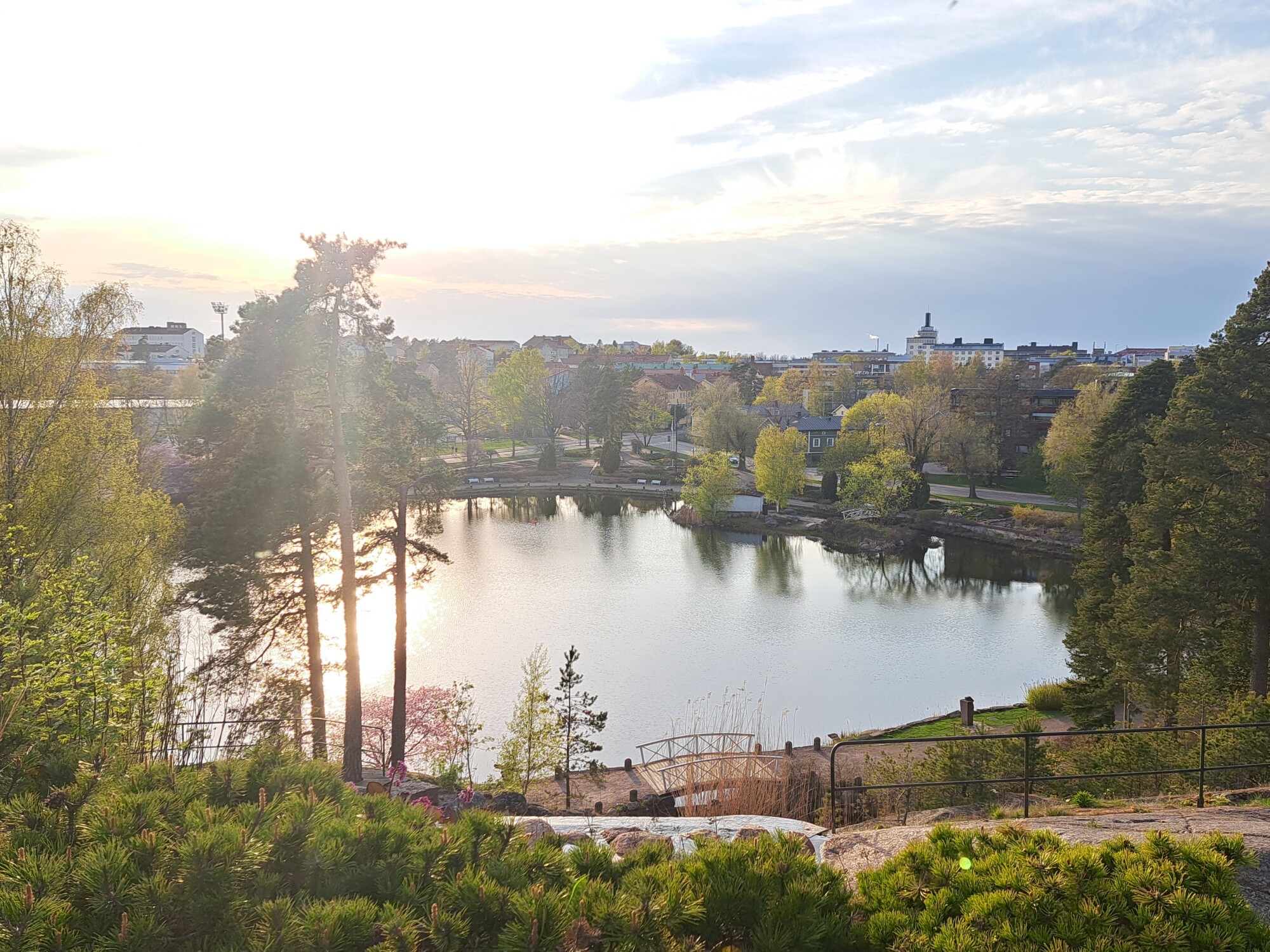 Seesteinen lampi puiden ja vehreyden ympäröimänä, auringonvalo paistaa korkeiden puiden läpi. Taustalla näkyy kaupungin rakennuksia ja taloja osittain pilvisen taivaan alla.