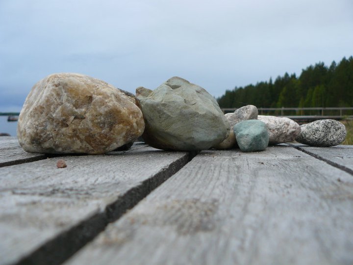 Useat erikokoiset ja -väriset kivet lepäävät vanhentuneella puisella laiturilla, ja taustalla näkyy järvenrantaa, puita ja pilvinen taivas. Kuva on otettu matalasta kulmasta.