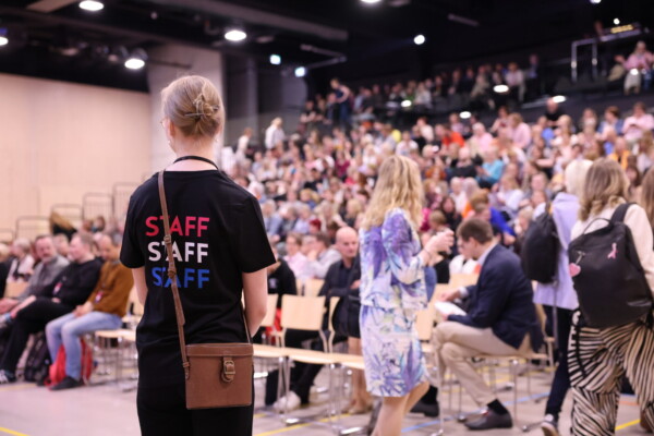 Henkilökunnan jäsen seisoo vastapäätä suurta istuvaa yleisöä kirkkaasti valaistussa sisätilassa. Henkilökunnan paidassa lukee punaisella, valkoisella ja sinisellä STAFF. Ihmiset keskustelevat ja asettuvat paikalleen ennen tapahtumaa.