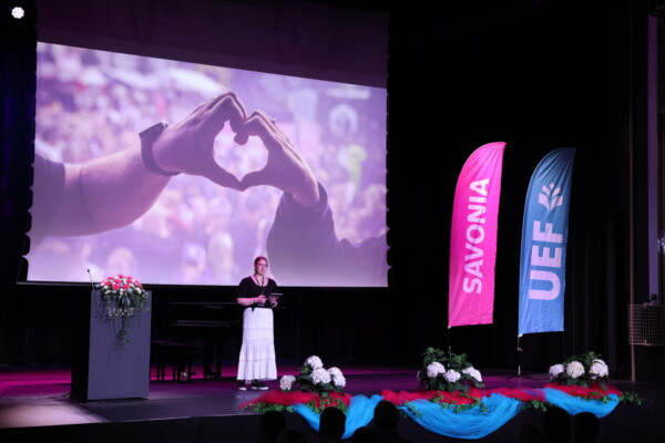 Nainen seisoo lavalla pitämässä puhetta kukkien koristaman korokkeen vieressä. Hänen takanaan on suuri näyttö, jossa näkyy kaksi kättä sydämen muotoisena. Oikealla näkyy kaksi värikästä lippua, joissa lukee SAVONIA ja UEF.