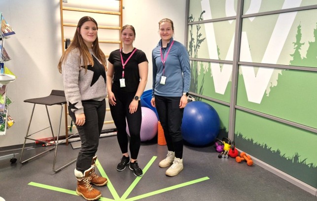 Kolme naista, joilla on nimikyltti, seisoo hymyillen kuntosalilla, jossa on jumppapalloja, käsipainoja ja kuntoilulaitteita. Heidän takanaan olevalla seinällä on vihreä luontoaiheinen kuvio.