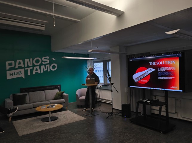 Henkilö seisoo puhujakorokkeella ja pitää esitystä yleisölle modernissa huoneessa, jonka seinä on vihreä PANOS HUB TAMO -seinä, jossa on harmaa sohva ja näyttö, jolla näkyy dia, jonka otsikko on THE SOLUTION.