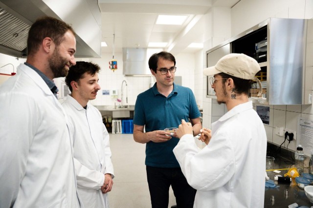 Neljä miestä seisoo laboratoriossa; kolmella on yllään valkoiset laboratoriotakit ja yhdellä sininen paita. He näyttävät keskustelevan keskenään: yksi heistä pitää kädessään laboratoriolaitteita, kun taas muut kuuntelevat tarkkaavaisesti.