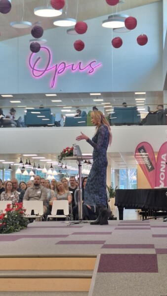 Nainen pitkässä sinisessä mekossa puhuu lavalla yleisölle modernissa, kirkkaasti valaistussa tapahtumatilassa, jonka taustalla on neon Opus-kyltti, värikäs ripustettu sisustus ja kaksi vaaleanpunaista mainosbanneria.