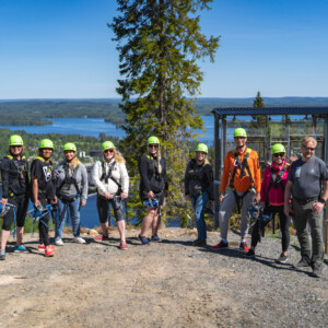 Ryhmä ihmisiä, joilla on kypärät ja valjaat, seisoo kukkulan laella, jonka taustalla on maisemallinen järvi ja metsä kirkkaan sinisen taivaan alla. He näyttävät olevan valmiita ulkoilmaseikkailuun tai laskuvarjohyppytoimintaan.