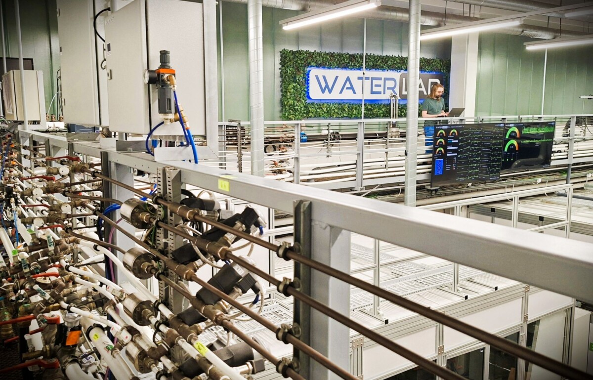Nykyaikainen laboratorio, jossa on putkia, mittareita ja mittareita metallirakenteen varrella, suuri näyttö, joka näyttää tietoja, ja henkilö, joka seisoo taustalla vihreällä seinällä olevan WATERLAB-kyltin vieressä.