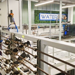 Nykyaikainen laboratorio, jossa on putkia, mittareita ja mittareita metallirakenteen varrella, suuri näyttö, joka näyttää tietoja, ja henkilö, joka seisoo taustalla vihreällä seinällä olevan WATERLAB-kyltin vieressä.