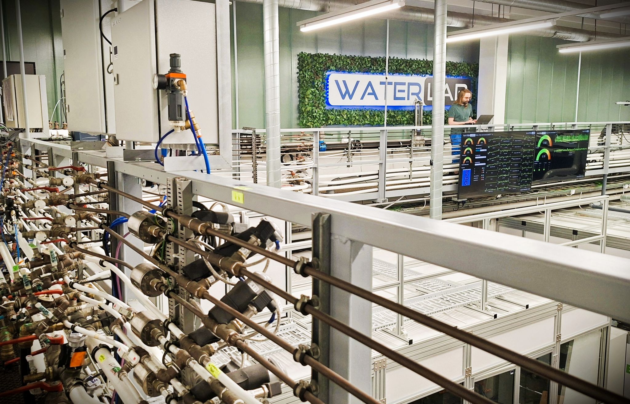 Nykyaikainen laboratorio, jossa on putkia, mittareita ja mittareita metallirakenteen varrella, suuri näyttö, joka näyttää tietoja, ja henkilö, joka seisoo taustalla vihreällä seinällä olevan WATERLAB-kyltin vieressä.