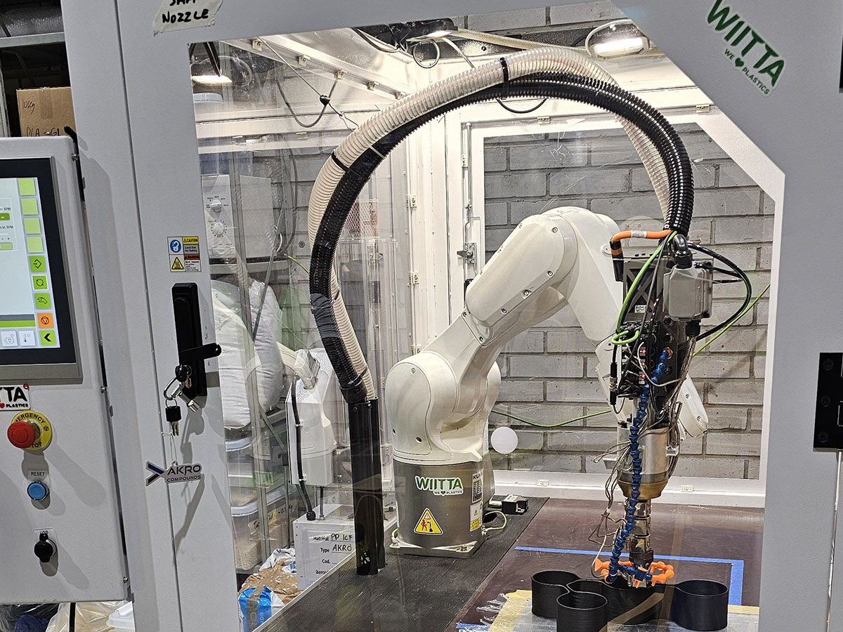 Suljetussa työtilassa oleva robottivarsi ohjaa kaapeleilla varustettua työkalua pinnan päällä. Lähellä on sylinterimäisiä esineitä, ohjauspaneeleja ja taustalla harmaa tiiliseinä.