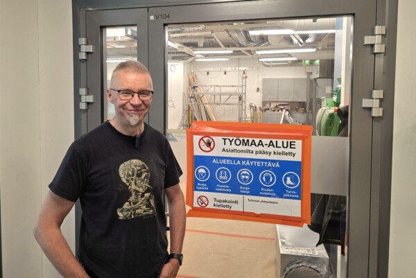 Lasit päässä oleva hymyilevä mies seisoo lasioven edessä, jossa on suuri suomeksi kirjoitettu turvallisuusmerkki, joka rajoittaa pääsyä rakennustyöalueelle ja luettelee vaadittavat suojavarusteet. Alue oven sisäpuolella näyttää työmaalta.
