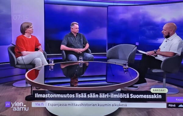 Kolme henkilöä istuu televisiostudion lavastuksessa keskustelemassa ilmastonmuutoksesta. Kaksi vierasta istuu pyöreän lasipöydän ääressä kaljupäistä juontajaa vastapäätä. Ruudulla näkyy suomenkielistä tekstiä ja uutisnauha, jossa on violetteja ja keltaisia graafisia elementtejä.