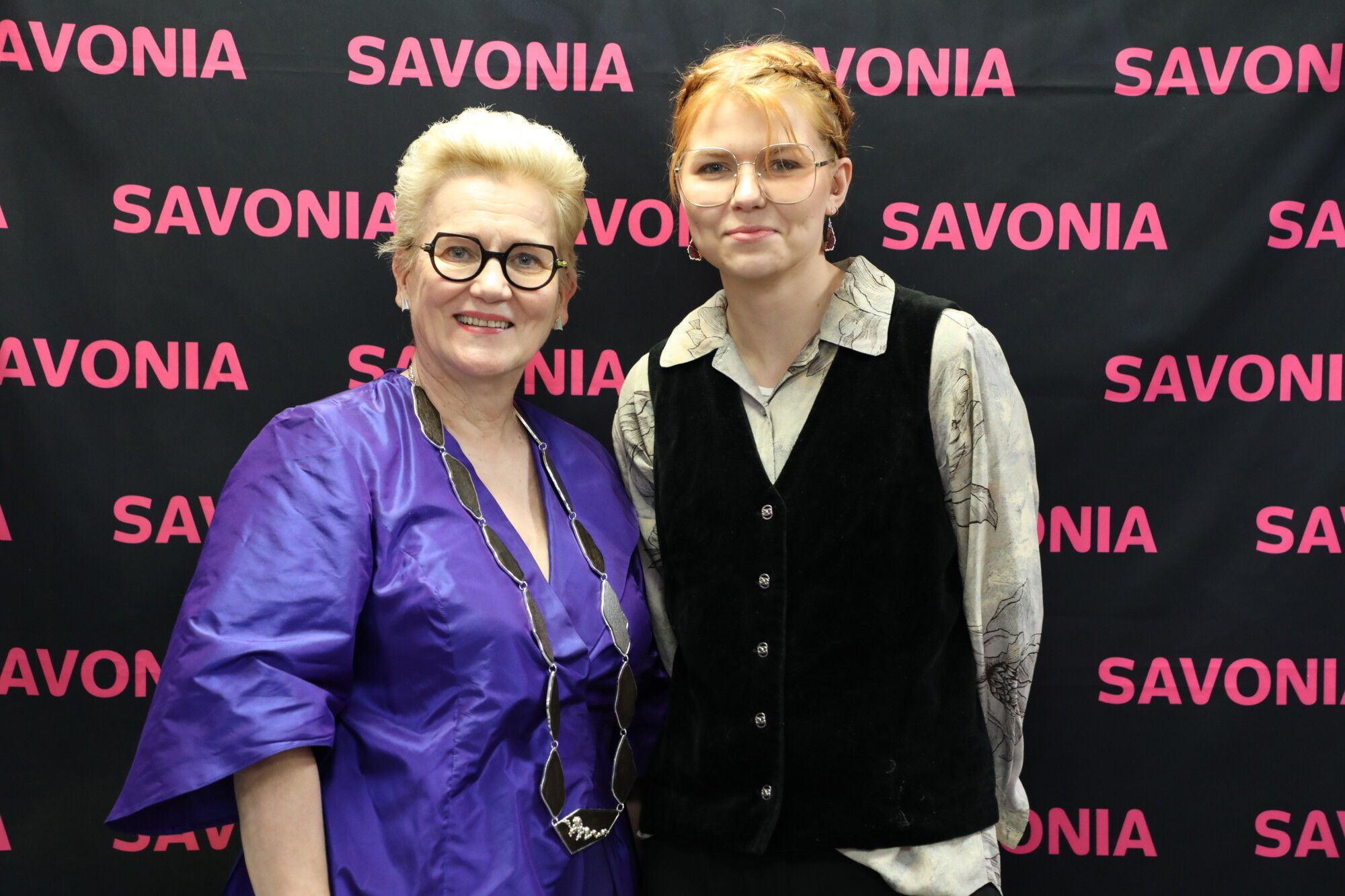 Kaksi henkilöä seisoo hymyillen taustakuvan edessä, jossa sana SAVONIA toistuu vaaleanpunaisilla kirjaimilla mustalla pohjalla. Toisella on yllään violetti mekko ja silmälasit; toisella on musta liivi, vaalea paita ja silmälasit.