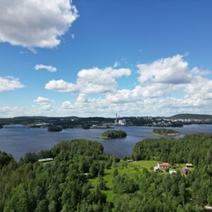 Maisemallinen ilmakuva järvestä, jossa on pieniä metsäisiä saaria, reheviä vihreitä puita ja hajallaan olevia taloja sinisen taivaan alla, jota koristavat pörröiset valkoiset pilvet. Horisontissa näkyy kaukana sijaitseva kaupunki ja kukkuloita.