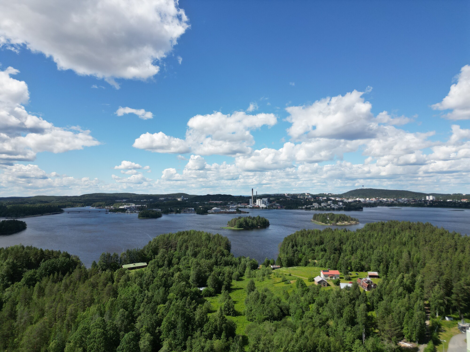 Maisemallinen ilmakuva järvestä, jossa on pieniä metsäisiä saaria, reheviä vihreitä puita ja hajallaan olevia taloja sinisen taivaan alla, jota koristavat pörröiset valkoiset pilvet. Horisontissa näkyy kaukana sijaitseva kaupunki ja kukkuloita.