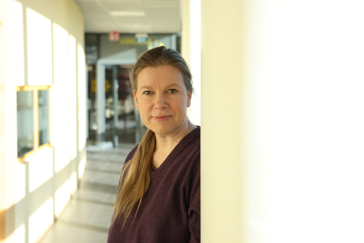 Nainen, jolla on pitkät, vaaleanruskeat hiukset poninhännässä, seisoo sisätilassa nojautuneena keltaista seinää vasten. Hänellä on yllään violetti villapaita, ja hän katsoo kameraan. Auringonvalo tulvii ikkunoista hänen takanaan.