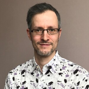 Mies, jolla on lyhyet tummat hiukset, silmälasit ja siistitty parta, hymyilee hieman. Hänellä on yllään valkoinen paita, jossa on mustia ja violetteja roiskeita, ja hän seisoo yksinkertaisen beige-värisen seinän edessä.
