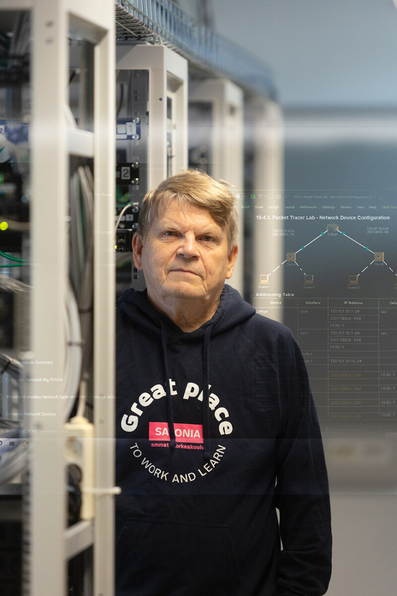 Vanhempi mies, jolla on yllään ”Great place to work and learn” -huppari, seisoo palvelinhuoneessa, ympärillään tietokonelaitteita ja taustalla näkyviä digitaalisia verkkokaavioita.