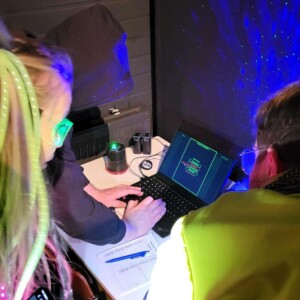Kolme henkilöä, joilla on yllään neonvärisiä asusteita ja heijastinliivejä, on kokoontunut kannettavan tietokoneen ympärille pimeässä huoneessa, jota valaisevat siniset ja vihreät valot. He tutkivat näytöllä näkyvää digitaalista koodia ja tekevät muistiinpanoja paperille.