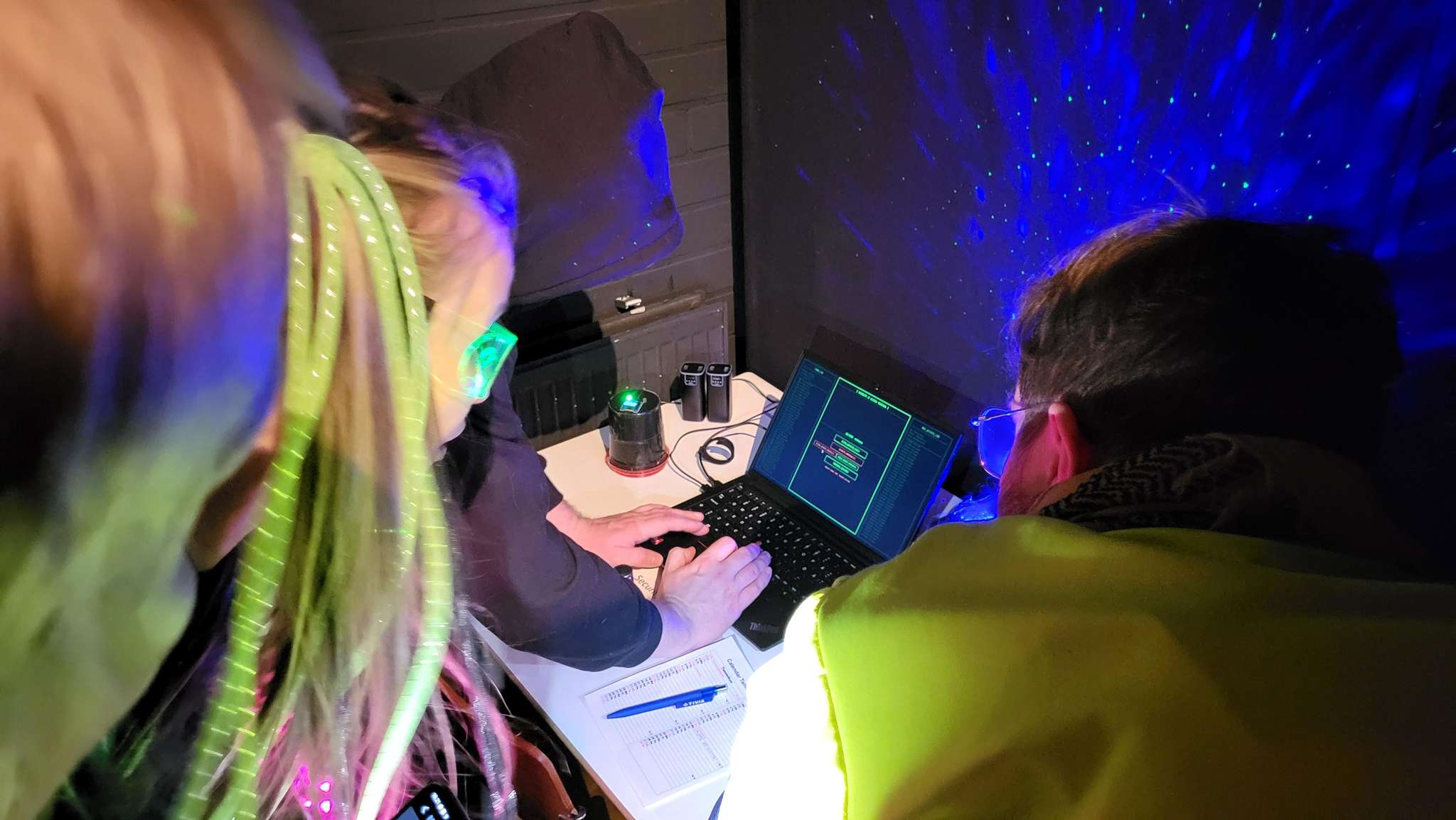 Kolme henkilöä, joilla on yllään neonvärisiä asusteita ja heijastinliivejä, on kokoontunut kannettavan tietokoneen ympärille pimeässä huoneessa, jota valaisevat siniset ja vihreät valot. He tutkivat näytöllä näkyvää digitaalista koodia ja tekevät muistiinpanoja paperille.