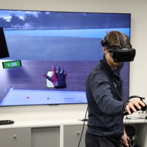 Henkilö, jolla on päässään VR-lasit ja kädessään ohjain, pelaa virtuaalista golfpeliä, joka näkyy hänen takanaan olevalla suurella näytöllä. Näytöllä näkyy golfkäsine, ajastin ja kuuden parhaan pelaajan tulokset.