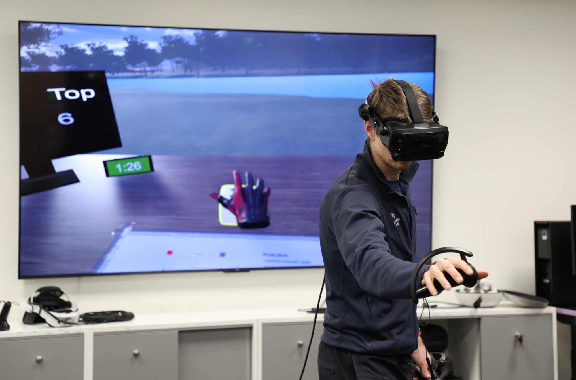 Henkilö, jolla on päässään VR-lasit ja kädessään ohjain, pelaa virtuaalista golfpeliä, joka näkyy hänen takanaan olevalla suurella näytöllä. Näytöllä näkyy golfkäsine, ajastin ja kuuden parhaan pelaajan tulokset.